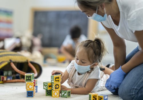 Teacher with mask talking with child with mask playing with blocks at a pre-school