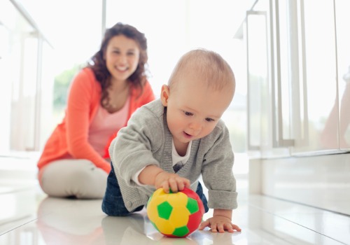 A mother watches her baby play with a ball, which is a great way to reach developmental milestones for a baby. If you're asking What are Gross Motor Skills, this is just one of many ways to build those skills.