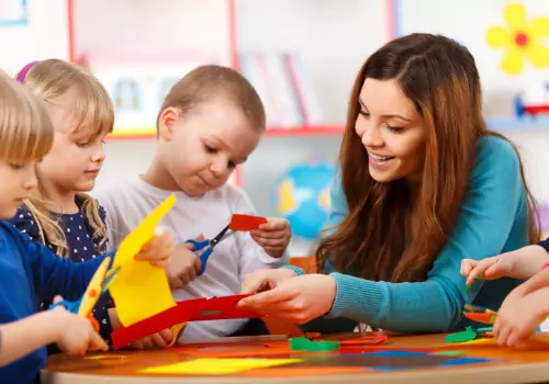 A teacher works on cutting paper with preschoolers. Modeling behaviors with enthusiasm is one of the tenants of Don Holdaway’s Foundations of Literacy