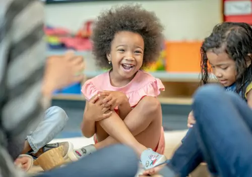 A preschooler laughs while sitting in a circle as part of daily routines, which is a part of the learning at daycare. What Does Daycare Teach My Child? It teaches how to be social.