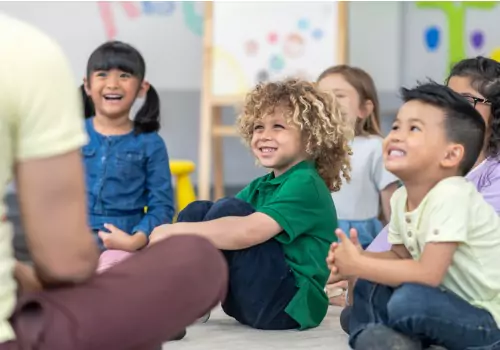 A group of of toddlers smile at their teacher as they sit listening to her. If you're asking What Does Daycare Teach My Child, PALS helps your children build confidence and learn.