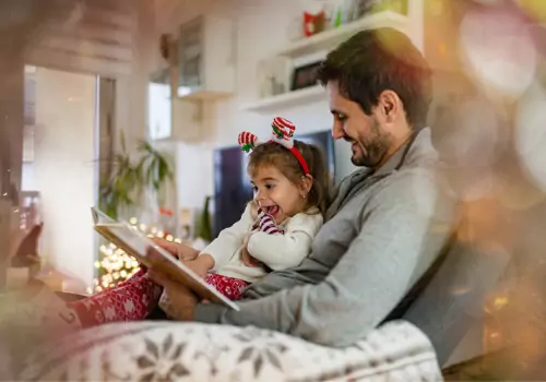 A dad reads to his toddler. Maintaining a routine is one of the easiest ways you can work on Keeping Kids Healthy During the Holidays.